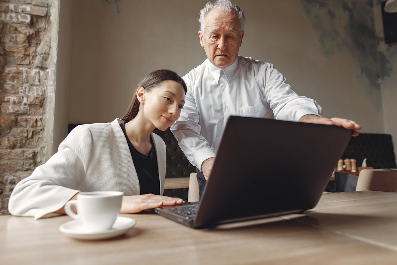 Low angle of positive man and woman of different ages in elegant outfit working remotely together with laptop and cup of coffee in modern workspace in loft style